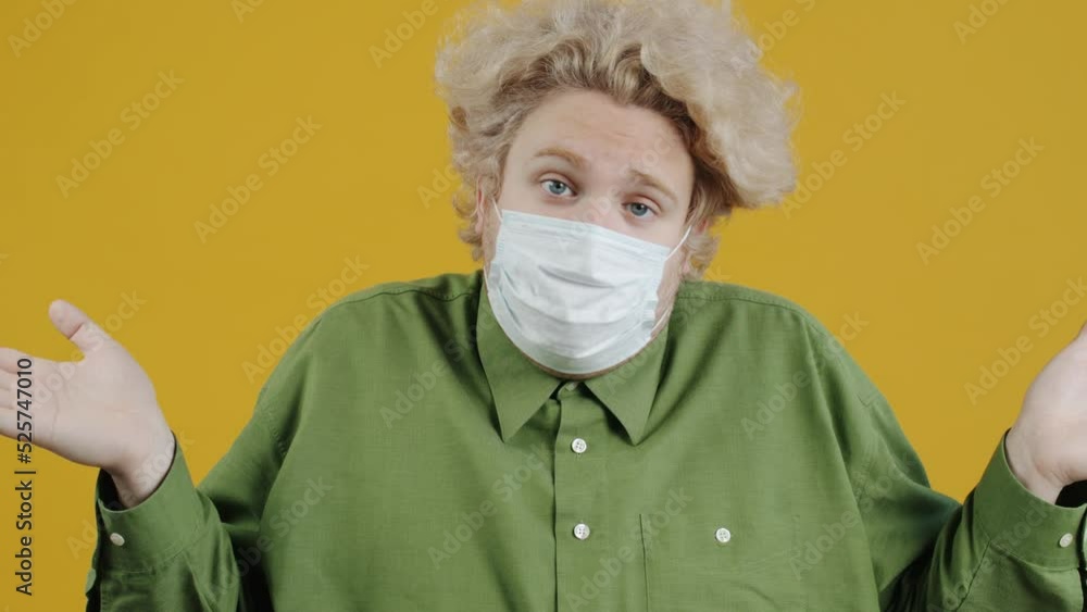 Portrait of tired and unhappy young man wearing medical face mask stretching arms feeling insecure on yellow background. People and pandemic concept. Portrait of tired and unhappy young man wearing medical face mask stretching arms feeling insecure on yellow background. People and pandemic concept.