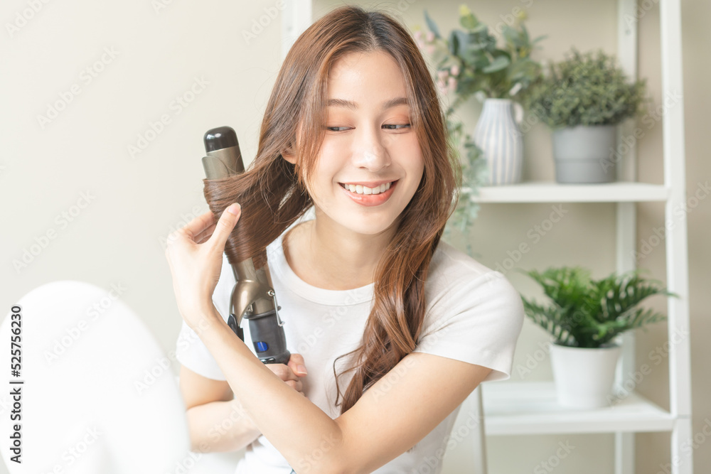 Beautiful curly brunette, attractive asian young woman, girl smiling using curler, curling iron long hair with ceramic curler straightener at home. Hairstyle and Hairdressing. Beauty and hair care . Beautiful curly brunette, attractive asian young woman, girl smiling using curler, curling iron long hair with ceramic curler straightener at home. Hairstyle and Hairdressing. Beauty and hair care .