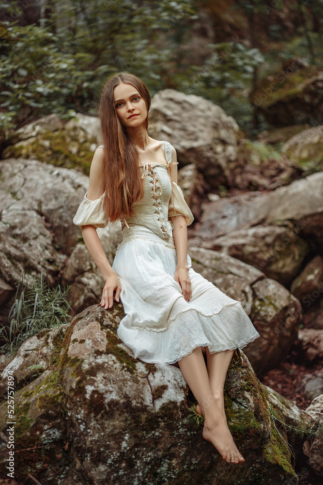 A girl in white sits on the waves in a wild forest. A girl in a corset sits on a stone in the forest.
