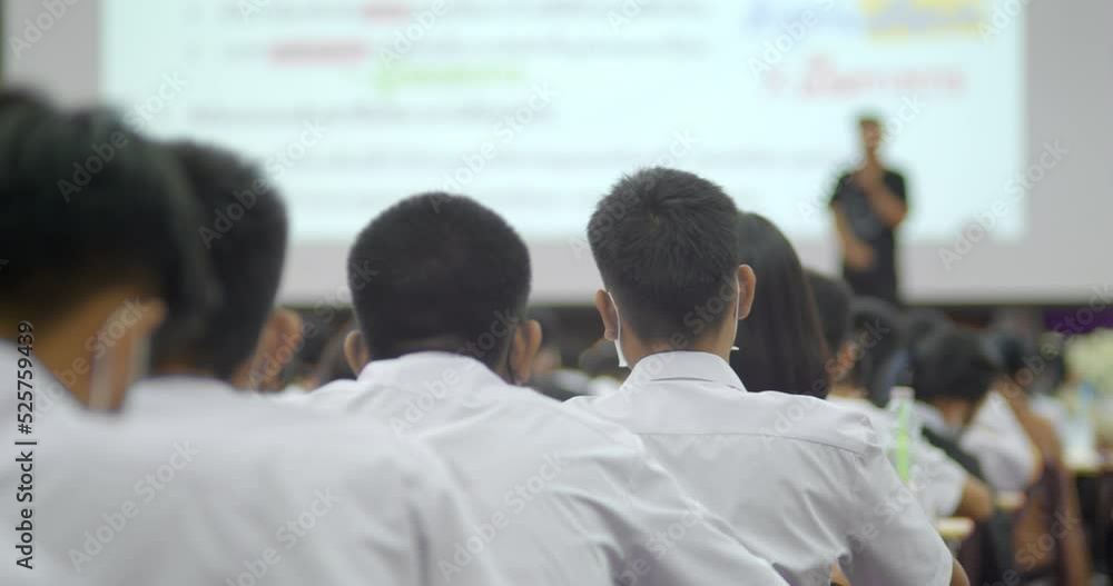 Slow motion scene, students in white uniforms are sitting in lecture halls in a large auditorium and raise their hands to answer the questions. A teacher stands in front of a large projector screen. Slow motion scene, students in white uniforms are sitting in lecture halls in a large auditorium and raise their hands to answer the questions. A teacher stands in front of a large projector screen.