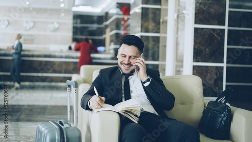 Pan shot of cheerful confident businessman talking mobile phone smiling and writing notes in notepad while sitting on armchair in hotel reception. Travel, business and people concept