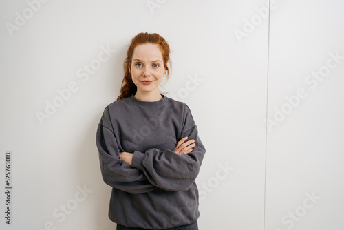 young redhead woman standing with crossed arms in front of white wall photo