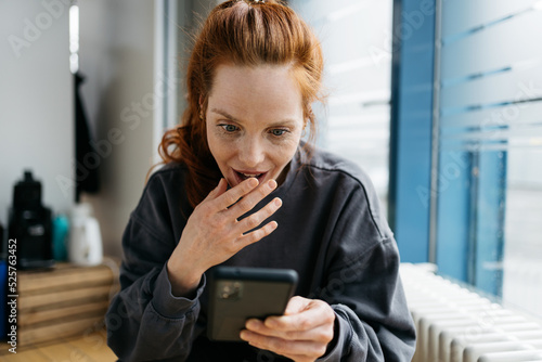 Картината върху платно young redhead woman looks at her cell phone in amazement