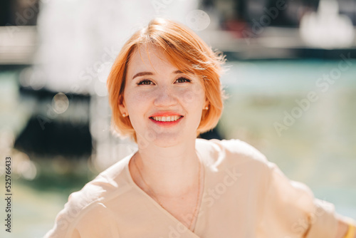 Young beautiful happy smiling woman with red short hair sitting outdoors in the city park near the fountain. Summer sunny day
