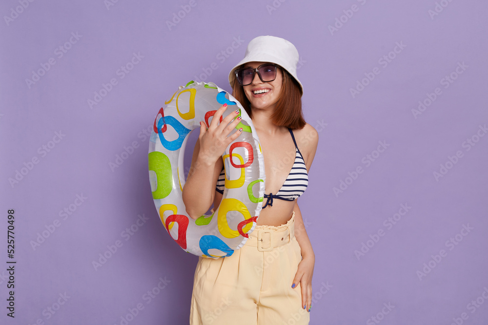 Fototapeta premium Portrait of pretty satisfied woman with rubber ring wearing panama and swimming suit posing isolated over purple background, looking away with toothy smile, enjoying her vacation.