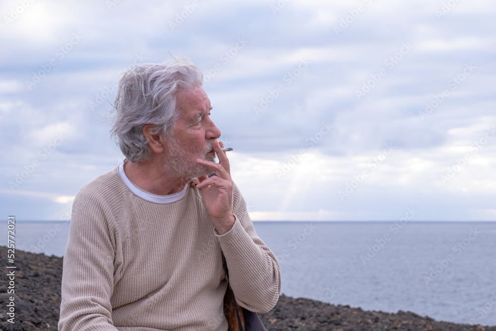 Black and white portrait of senior man sitting on the cliff in sea excursion smoking a cigarette looking at the horizon. White-haired grandfather enjoying vacation and freedom