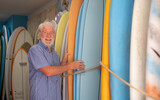 Happy senior caucasian man choosing a surfboard in a shop selecting from different sizes and color