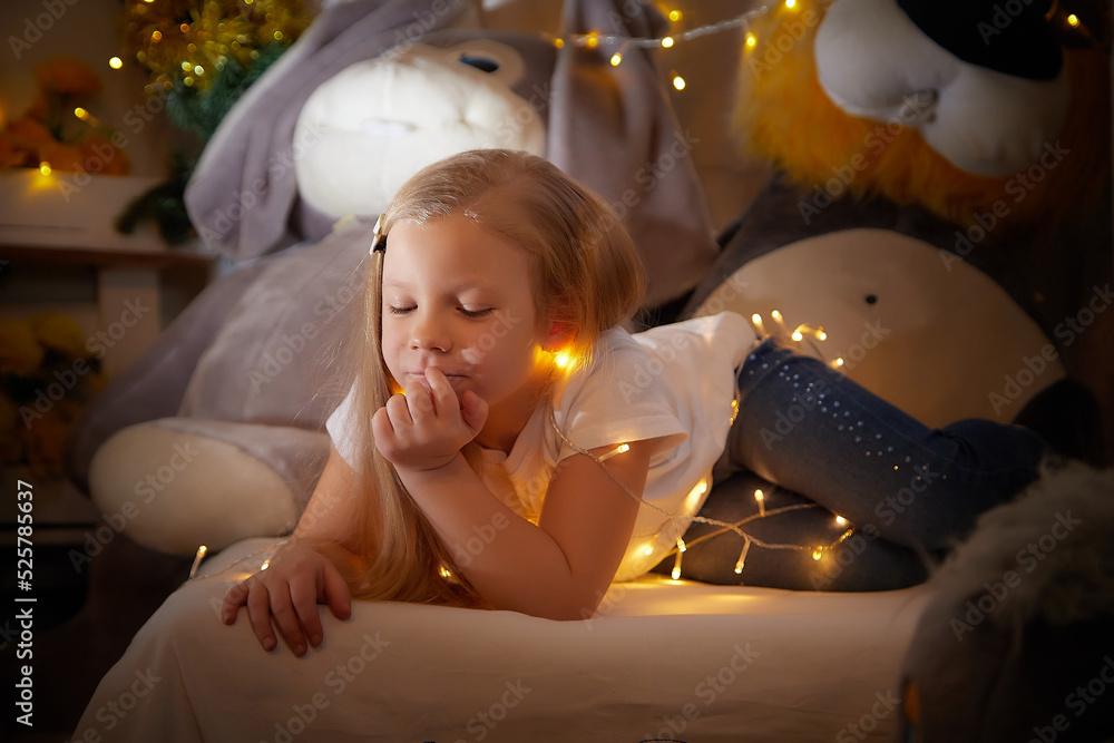 Cozy girl child with a garland on the bed in the children room, looking at the lights in night or evening of Christmas or New Year Cozy girl child with a garland on the bed in the children room, looking at the lights in night or evening of Christmas or New Year