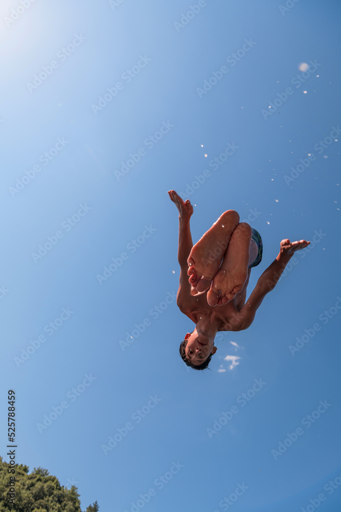 Young teen boy jumping flying and diving in the river. Clear blue sky and trees in distance as a natural background.