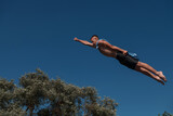 Young teen boy wearing a towel as a superhero scarf flying and diving in river. Clear blue sky and trees in distance as a natural background.