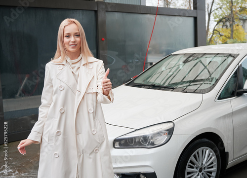 Portrait of young beautiful woman near her white modern car at carwash station outdoor. Female owner smiling to the camera, showing thumbs up.