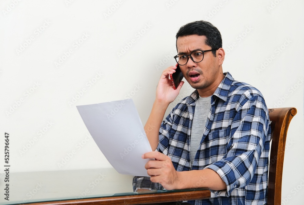 Asian man showing shocked expression when on a phone call and read letter Asian man showing shocked expression when on a phone call and read letter