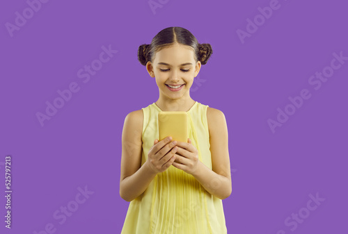 Smiling cute adorable preteen girl enjoys using modern smartphone isolated on purple background. Happy caucasian little girl looking at cellphone screen. Modern devices and children concept.