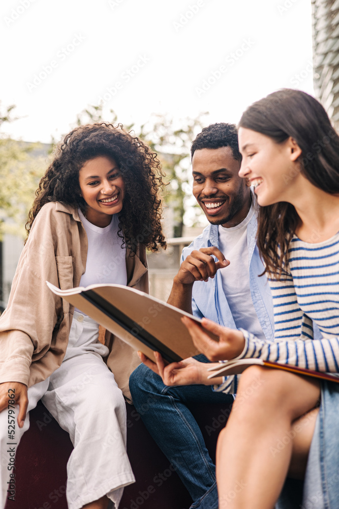 Group of young interracial students discussing homework looking at exercise book sitting on campus. Brunette guy and girls wear spring casual clothes. Education concept - obrazy, fototapety, plakaty 