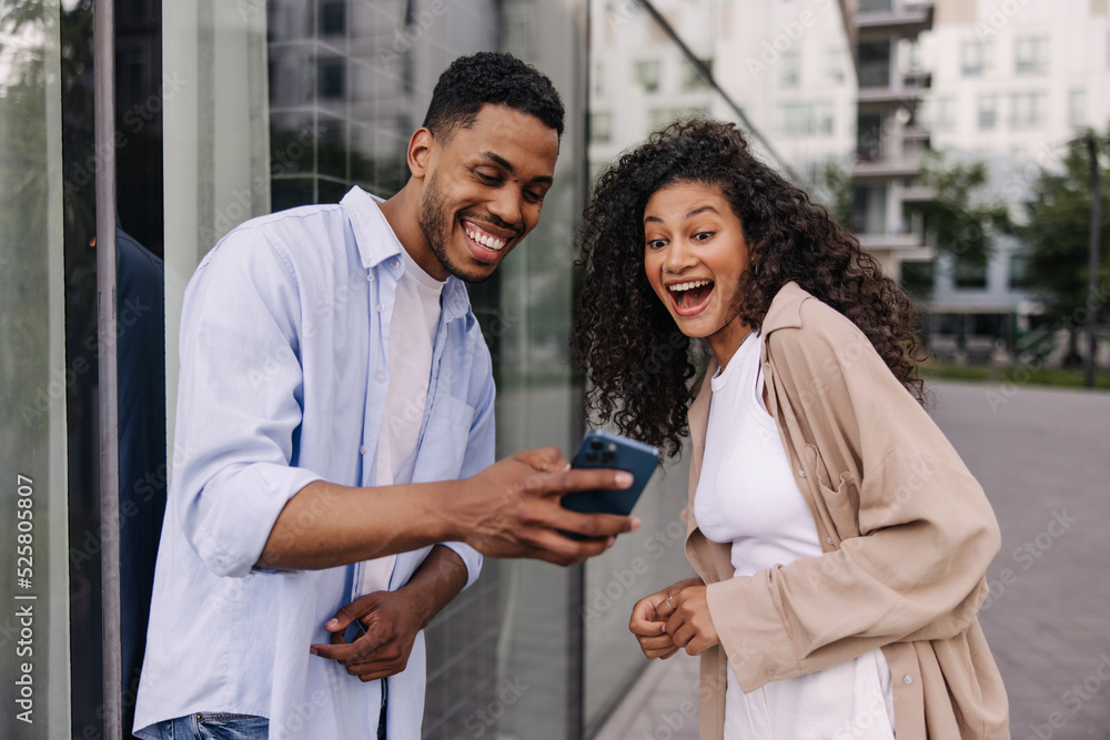 Joyful young african students are watching funny video on phone standing on street. Brunette guy and girl wear casual clothes. Technology concept Joyful young african students are watching funny video on phone standing on street. Brunette guy and girl wear casual clothes. Technology concept