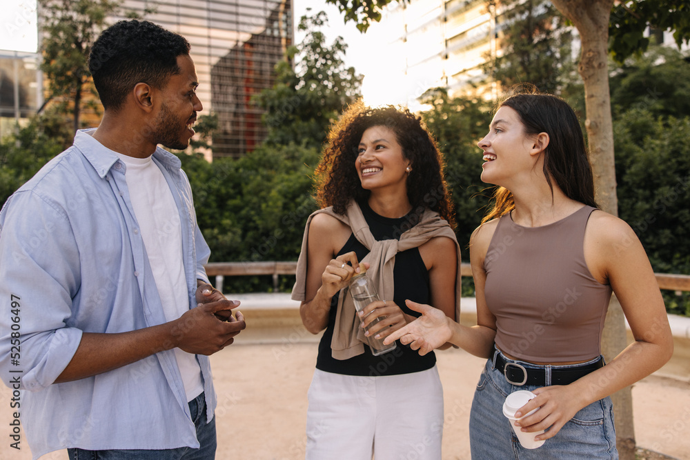 Obraz premium Cheerful young interracial friends discuss various topics strolling through city during day. Brunette guy and girls wear casual clothes. Relaxed lifestyle, concept