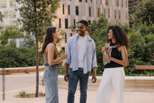 Cheerful young caucasian, african friends chatting and walking outdoors during day. Guy and girls wear casual clothes in spring. Happy day concept