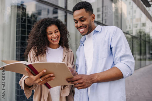 Happy young african guy and girl read notes in notebook standing outdoors. Brunette students wearing casual clothes are preparing for seminar. Study concept