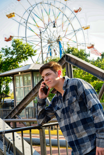 Handsome young man standing in front of iron railing and talking on a cellphone with a ferris wheel in the background.