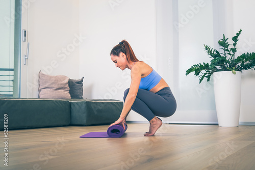 Young yoga woman rolling her violet mat after a yoga class on wooden floor near a window in her linving room photo