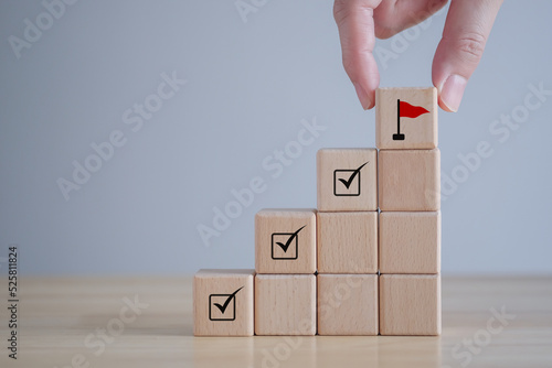 Checklist Survey and assessment. quality Control. Goals achievement and business success. Hand Holding wooden block with red flag icon and check mark on cube ladder.