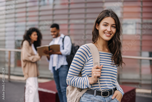 Selective focus on caucasian young girl with backpack looking at camera outdoors. Interracial university students posing near building. Lifestyle, different emotions, leisure concept