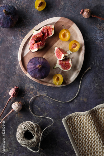 Autumn fruits, black fig and yellow plums on wooden tray. Flat lay, top view on dark textured background with dry poppy plants and hemp cord. Fruits in Fall season.