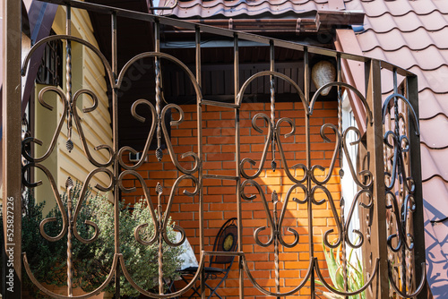 Summer veranda on second floor of country house. Fencing of terrace with curly lattice. Back wall is made of facing bricks. Garden furniture. Table and two chairs in far corner. Secluded relaxation.