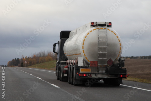Petrol transportation, white fuel tank truck with ADR sign 33 1203 on suburban empty asphalted road at autumn evening on gray sky background, flammable liquid goods transportation