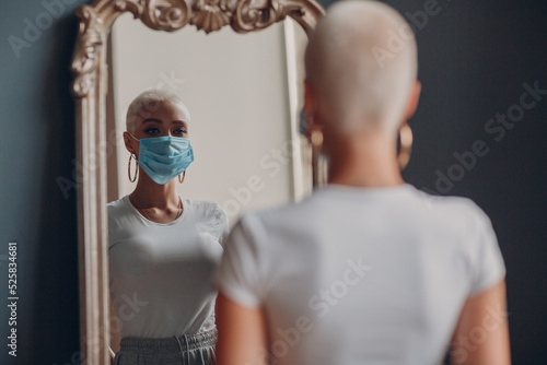 Millenial young woman in medical protective face mask with short blonde hair portrait standing at mirror indoor