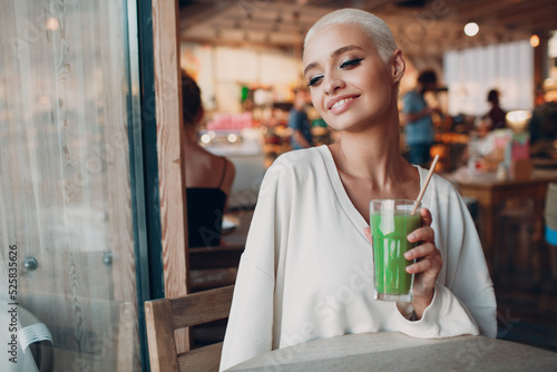 Portrait of young smiling millenial european short haired woman with green smoothie at cafe. Beautiful happy blonde girl indoor.