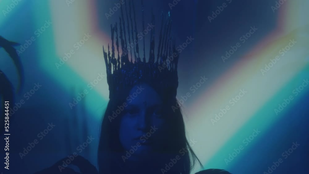 Young woman with face gem and gothic crown dressed in feather outfit standing in dark room with rainbow light projection Young woman with face gem and gothic crown dressed in feather outfit standing in dark room with rainbow light projection