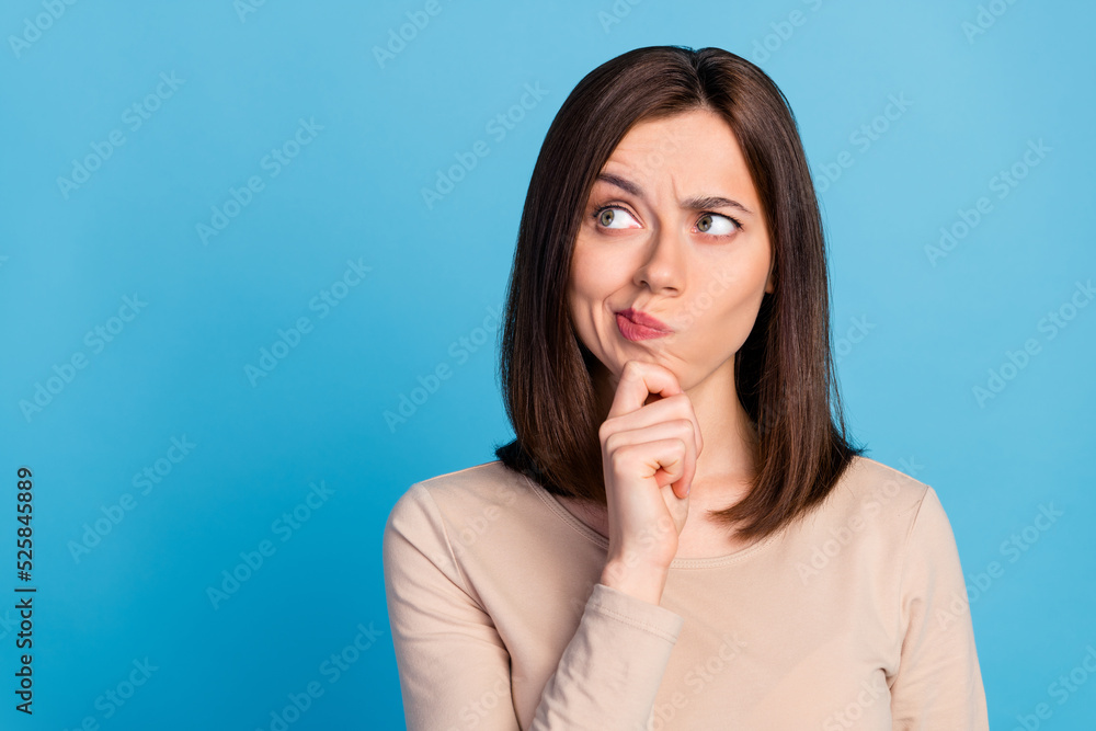 Photo of creative young lady hand touch chin look interested empty space isolated on blue color background Photo of creative young lady hand touch chin look interested empty space isolated on blue color background