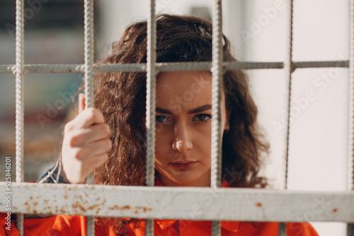 Foto Young brunette curly woman in orange suit behind jail bars