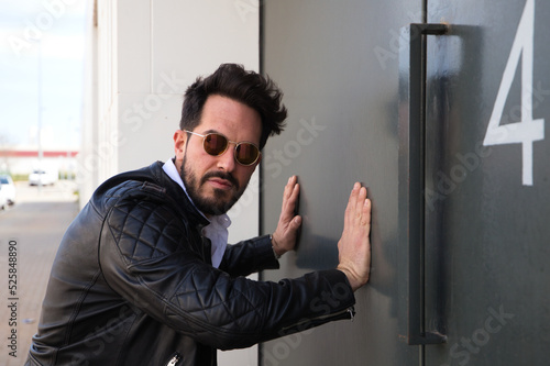 handsome young man with beard, sculpted body and sunglasses is leaning with both hands on the grey door of his garage looking at the camera for the portrait. Concept different expressions.