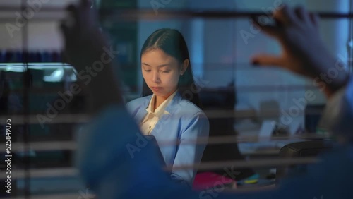 Shooting through glass of concentrated Asian woman messaging online on laptop with blurred boss peeping after employee and closing blinders. Corporate lifestyle concept