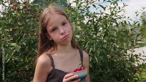 An offended, upset teenage girl is crying in the garden against the background of a cherry tree on a summer day, holding a bowl of cherries in her hands
