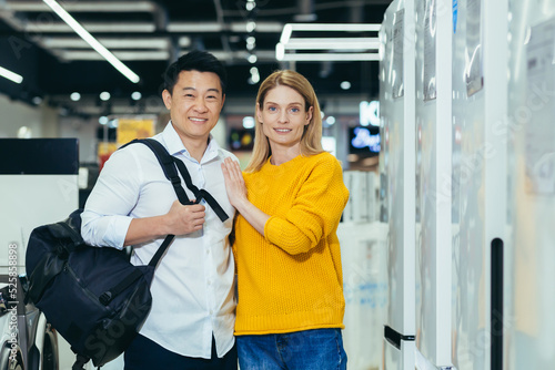 Portrait of a young married couple shopping in a home appliance supermarket, a diverse family of a man and a woman smiling and looking at the camera, choosing and buying home appliances © Liubomir