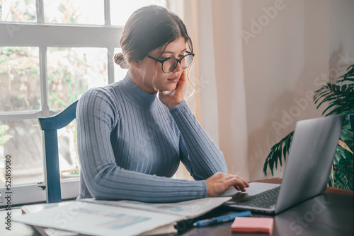 One young pretty woman studying and doing homework at home on the table. Female teenager using laptop or computer surfing the net indoor. photo