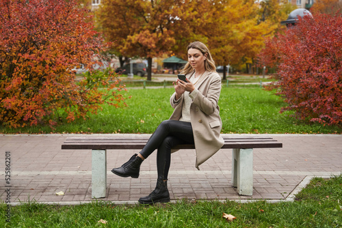 a beautiful woman against the backdrop of autumn trees in the park sits on a bench and looks at something in a smartphone. beautiful autumn portrait