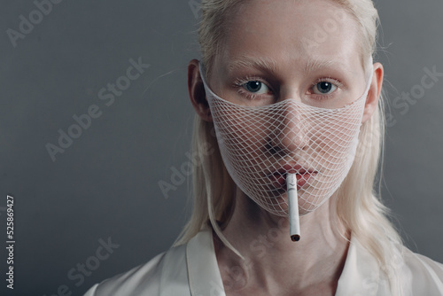 Closeup portrait of white caucasian albino blond woman fashion model wearing quarantine medical face mask coarse mesh net with cigarette on grey background