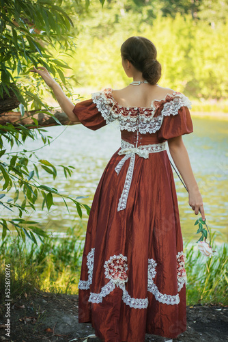 Beautiful young woman in 18-19 century dress in the forest by the pond. The romantic girl, view from the back.