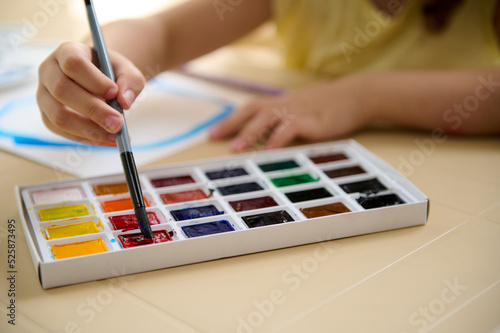 Details: small hand of a talented cute child, holding paintbrush and mixing colorful paints on a professional watercolor palette. Learning peinture. Art class. Kids education and hobby. Back to school