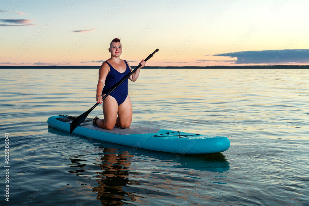 a woman in a closed swimsuit with a mohawk on her knees on a SUP board with an oar floats on the water against the background of the sunset sky.