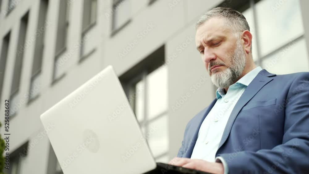 Mature gray-haired business man in formal suit working on laptop online remotely. Sitting on a bench near office building outside. senior employee uses a computer outdoors urban background city street Mature gray-haired business man in formal suit working on laptop online remotely. Sitting on a bench near office building outside. senior employee uses a computer outdoors urban background city street
