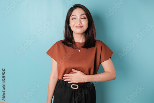 Pleased cheerful Asian woman keeps hand on belly feels full after delicious dinner stands against blue background.
