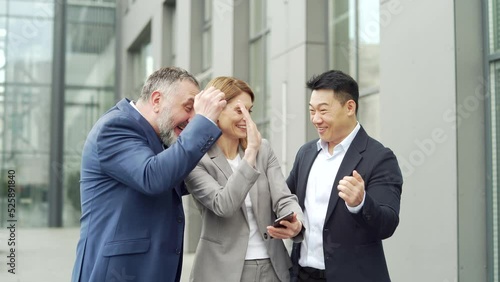 group of colleagues fellow employees read good news looking at smartphone. Happy team rejoices about successful investment while using, browsing mobile phone. Standing outside near an office building