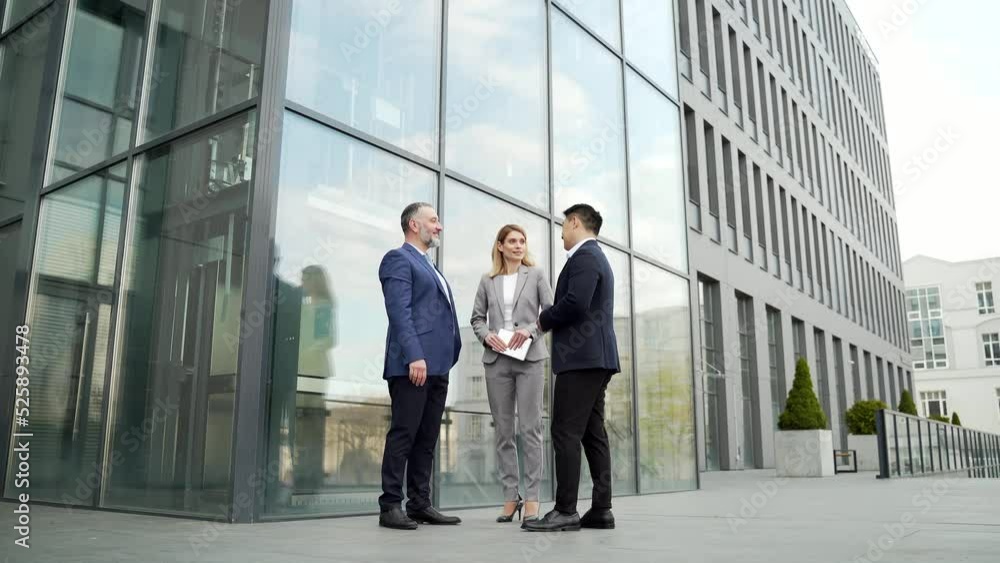 International business team meeting. mature man, Caucasian woman, Asian employee communicate . group of entrepreneurs in formal suits discuss a project document near office center on street. Outside International business team meeting. mature man, Caucasian woman, Asian employee communicate . group of entrepreneurs in formal suits discuss a project document near office center on street. Outside
