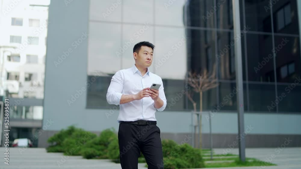 Successful asian business man walks down the city street background of urban office building using a smartphone. Confident businessman, male employee browsing, chatting, texting with phone in hands Successful asian business man walks down the city street background of urban office building using a smartphone. Confident businessman, male employee browsing, chatting, texting with phone in hands