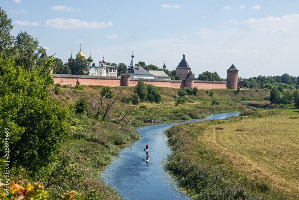 Panoramic view of the old brick walls of the Suzdal Kremlin on a high hill on the banks of the beautiful Kamenka river among the green grass on a sunny summer day in Russia